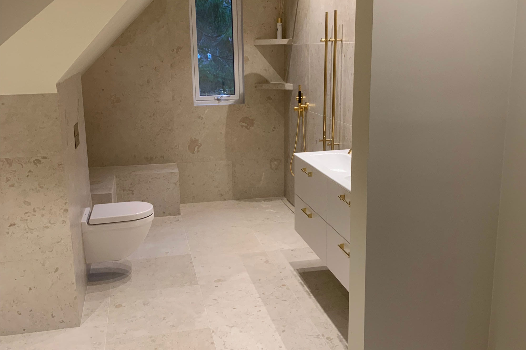 Modernt badrum med ljusa stenplattor och guldfärgade duschdetaljer